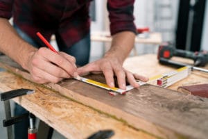 Eine Person in Werkstattumgebung misst und markiert ein Holzbrett mit einem Zollstock und einem roten Bleistift. Handwerkliches Arbeiten mit Fokus auf Genauigkeit.